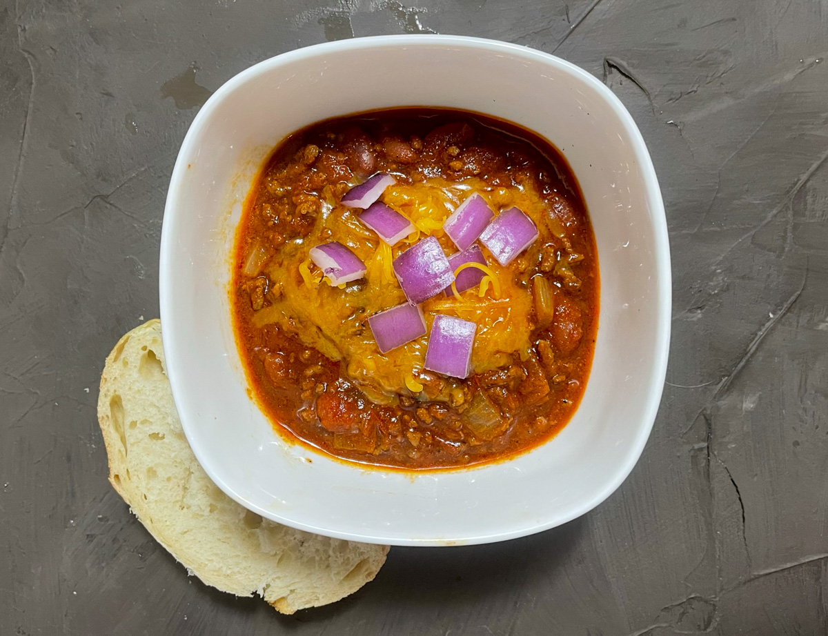Texas Roadhouse chili served in a bowl with cheddar cheese and onions along with a slice of bread