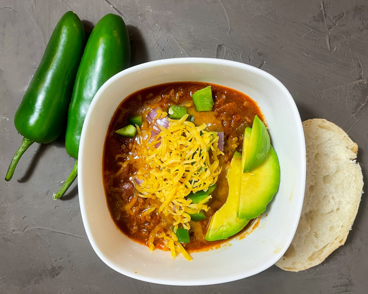 Texas chili with multiple toppings beside a piece of bread and two whole jalapeno peppers