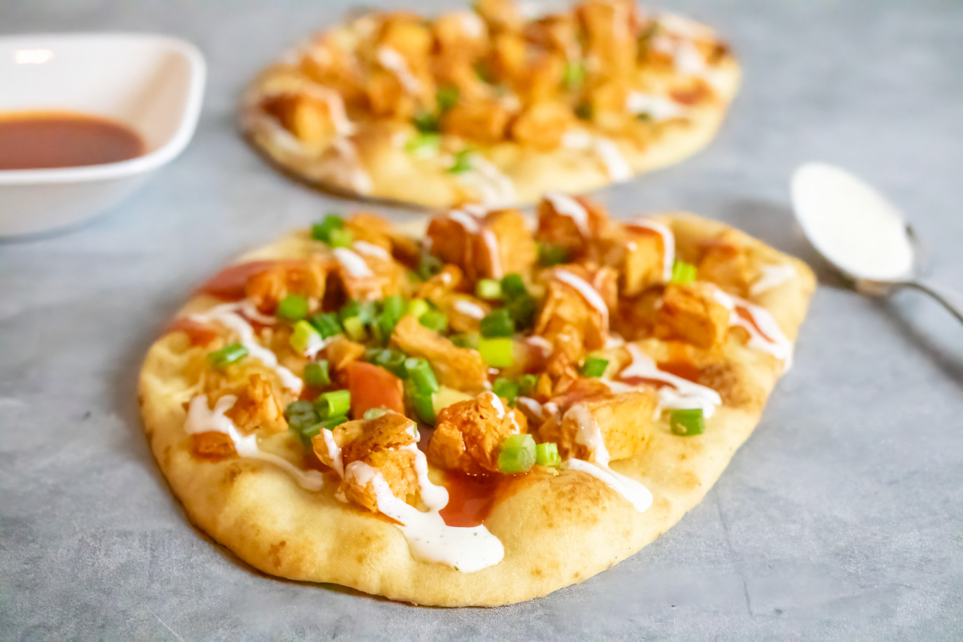 Two buffalo chicken flatbread pizzas, a bowl of buffalo sauce, and a spoon coated in ranch dressing