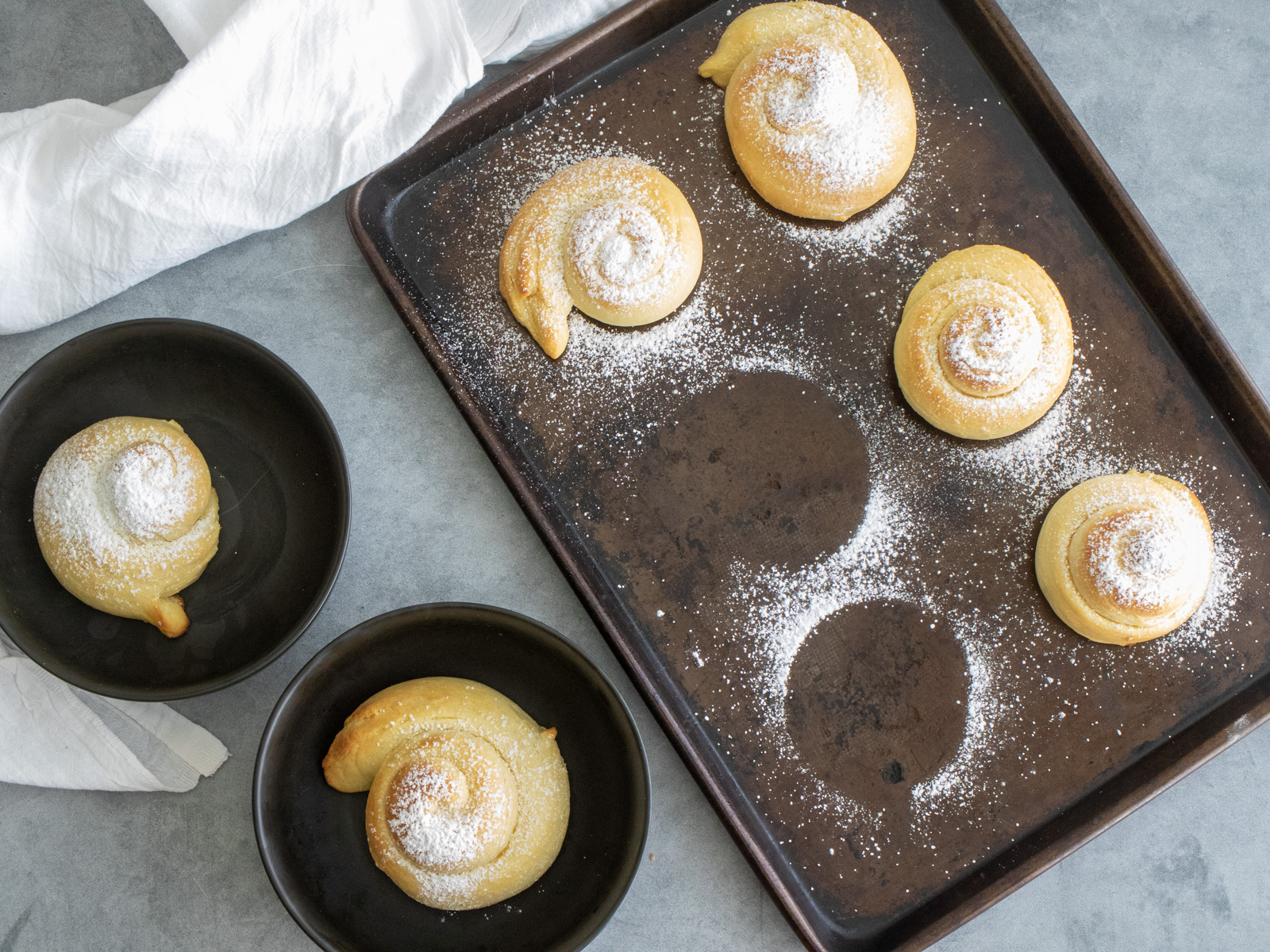 Pan de Mallorca rolls, covered with powdered sugar. 4 are still on baking sheet, 2 others in bowls, with a white towel beside.