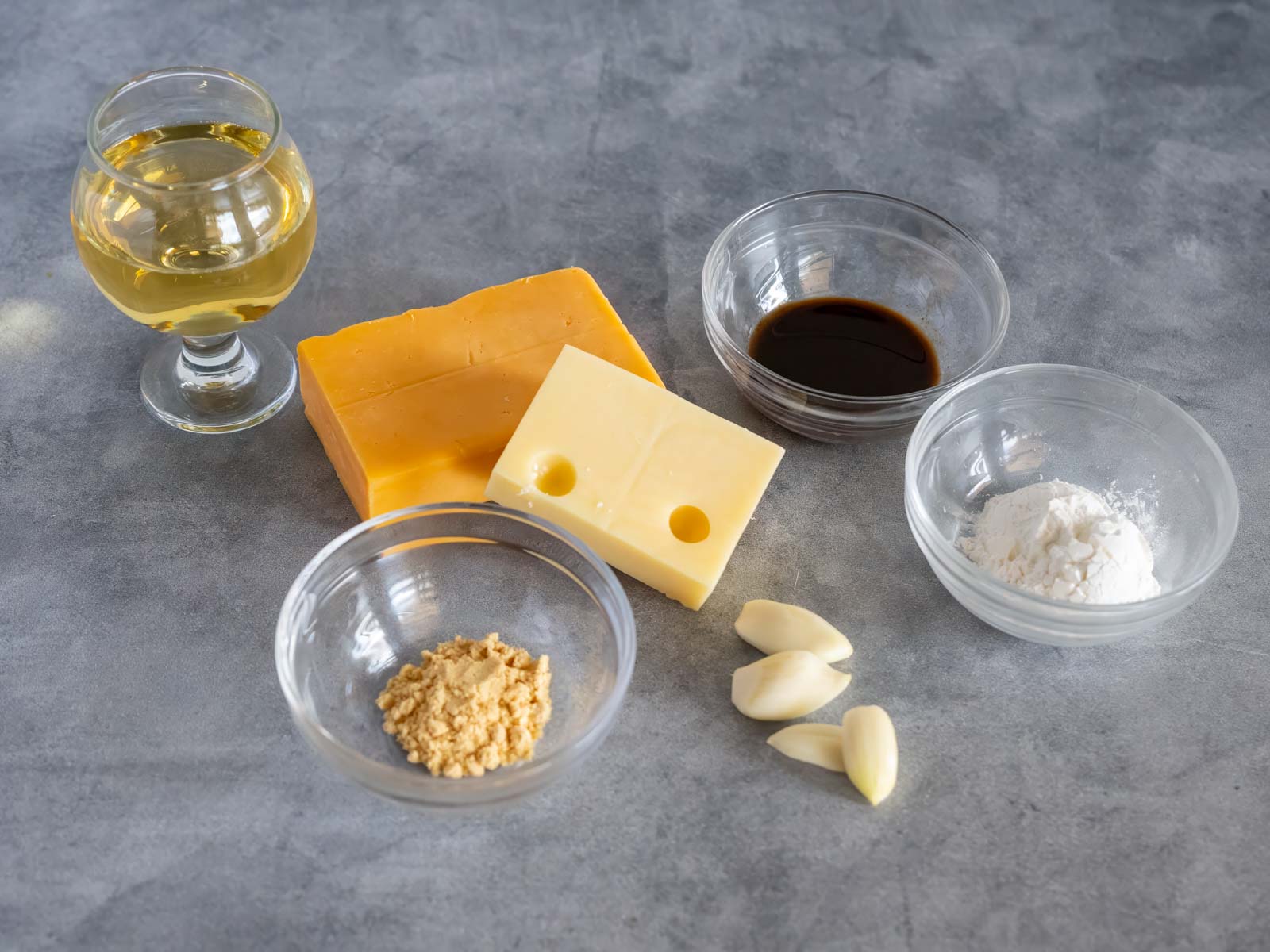 Melting Pot Wisconsin Cheedar Cheese fondue ingredients: cheddar and Swiss cheese blocks, garlic cloves, a snifter of light beer, and small bowls of dry mustard powder, Worcestershire sauce, and corn starch