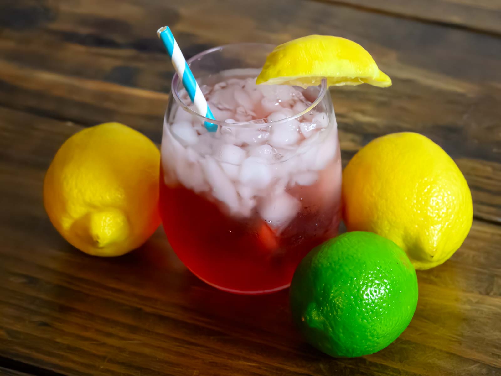 Cayman Lemonade in a glass over ice, on a wood table, surrounded by lemons and limes.