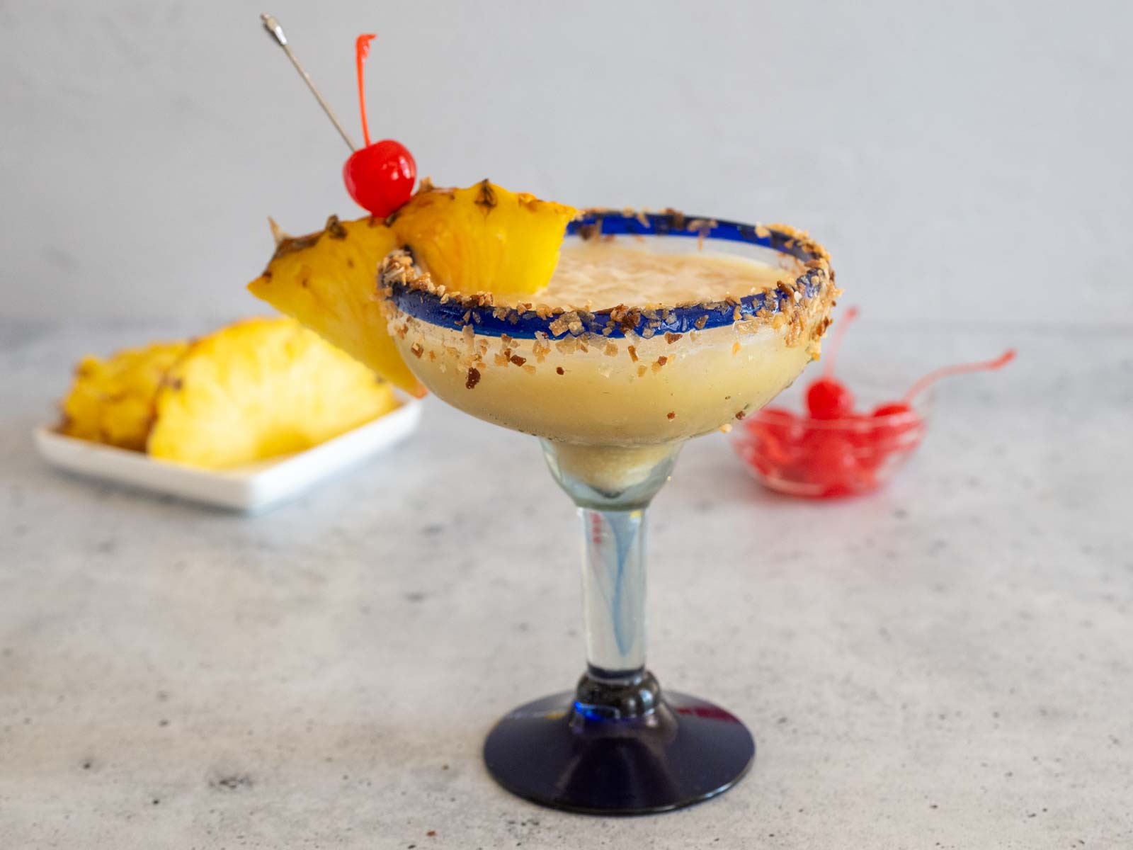 Cheddar's Painkiller Cocktail in a margarita glass, garnished with pineapple and a cherry, and with a straw. Dishes of extra pineapple and cherries are in the background.