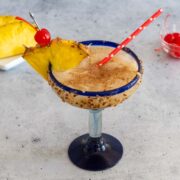 Cheddar's Painkiller Cocktail in a margarita glass, garnished with pineapple and a cherry, and with a straw. Dishes of extra pineapple and cherries are in the background.