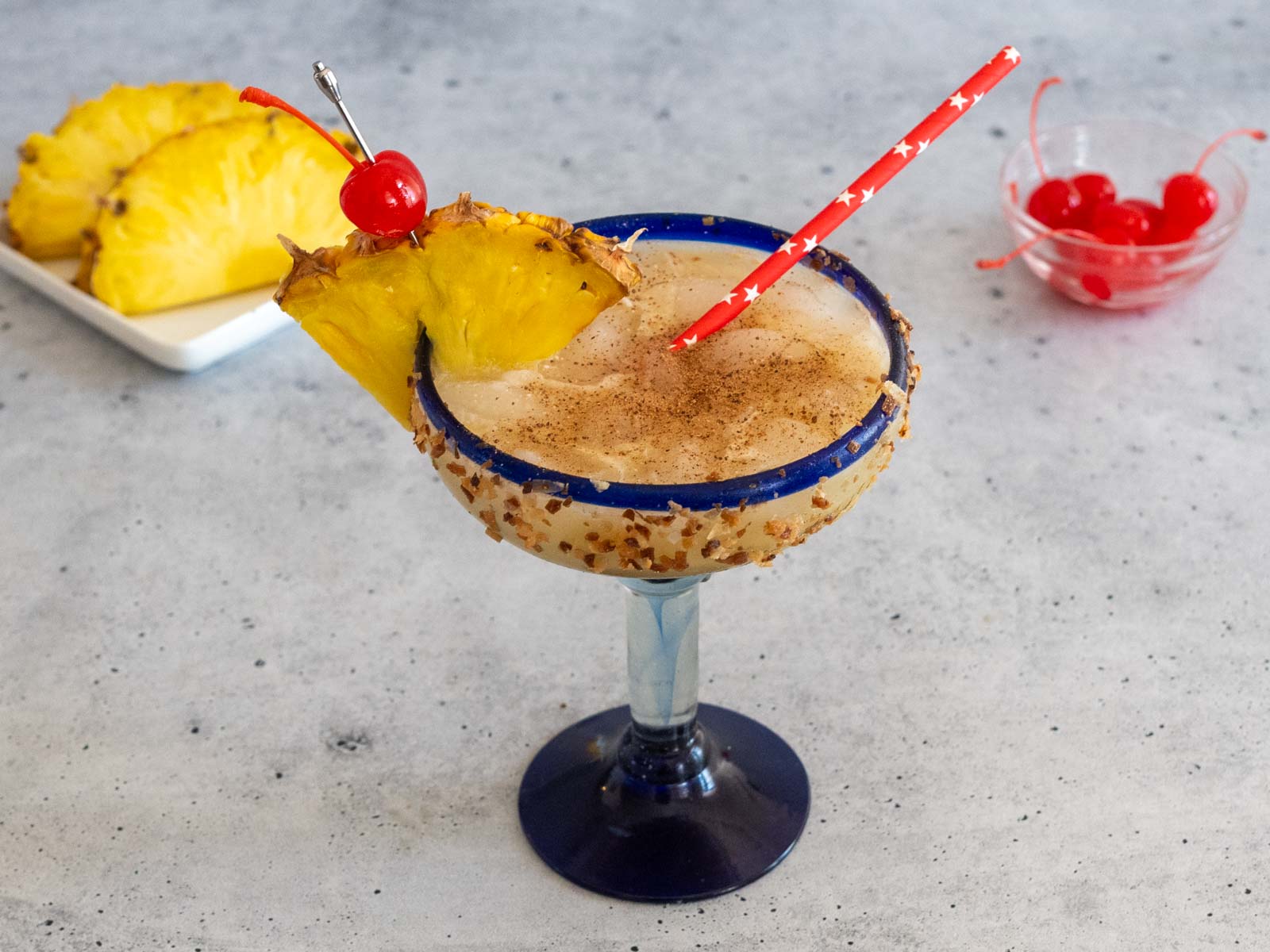 Cheddar's Painkiller Cocktail in a margarita glass, garnished with pineapple and a cherry, and with a straw. Dishes of extra pineapple and cherries are in the background.