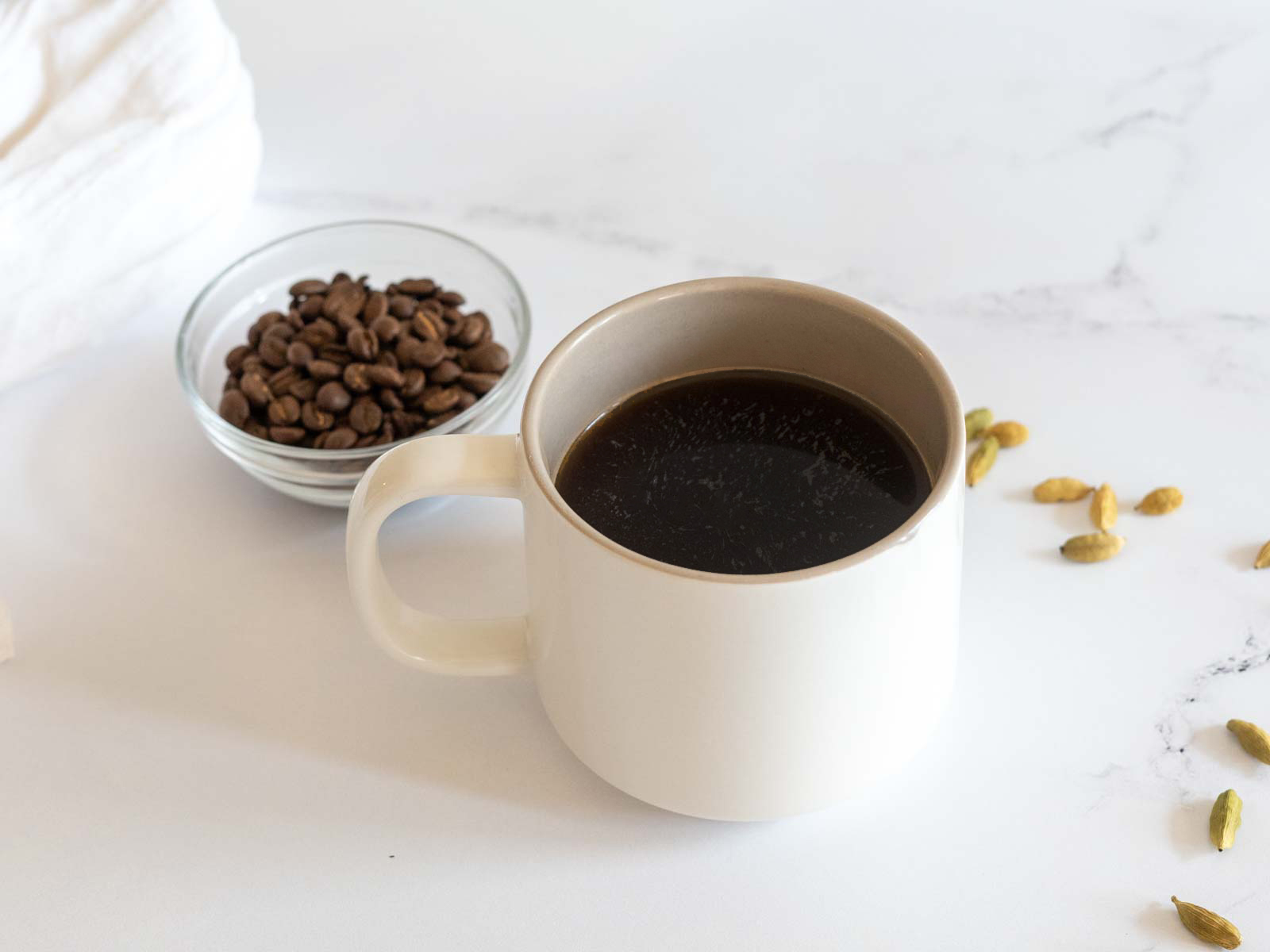 Cardamom coffee in a mug, with whole coffee beans and cardamom pods in the background